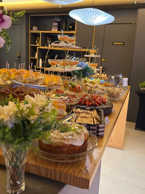 Vista da mesa Áurea com bolos, frios, frutas e decoração floral